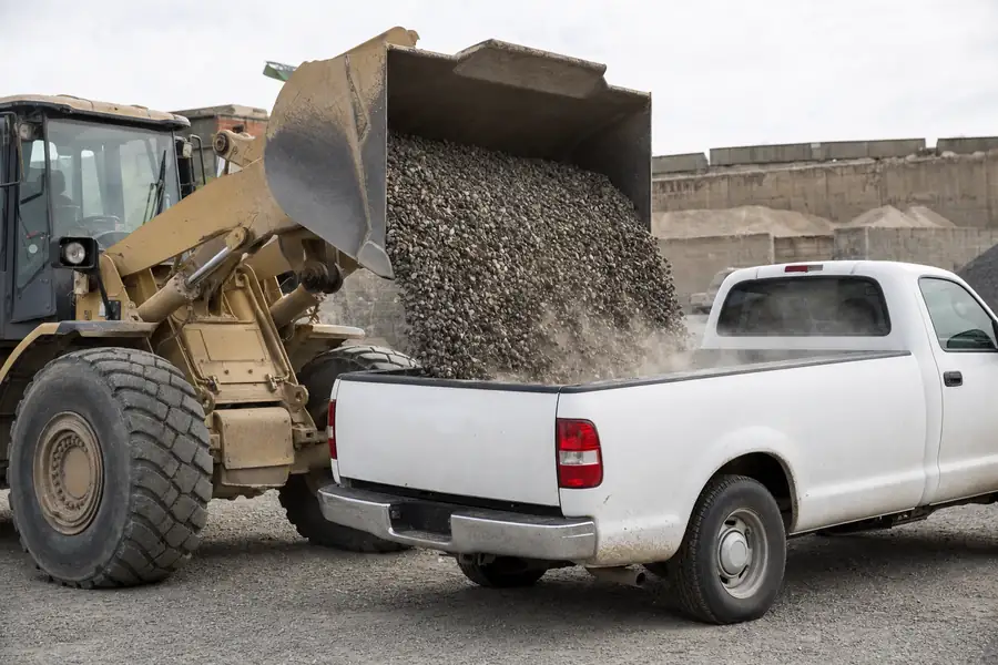 Action photo at a landscape supply yard: a wheel loader bucket releasing a full cubic yard of 3/4-inch gravel into the bed of a white half-ton pickup that visibly settles on its rear suspension, dust catching the sunlight, weighbridge and material bins in the background, candid working-yard atmosphere with realistic machine and stone detail