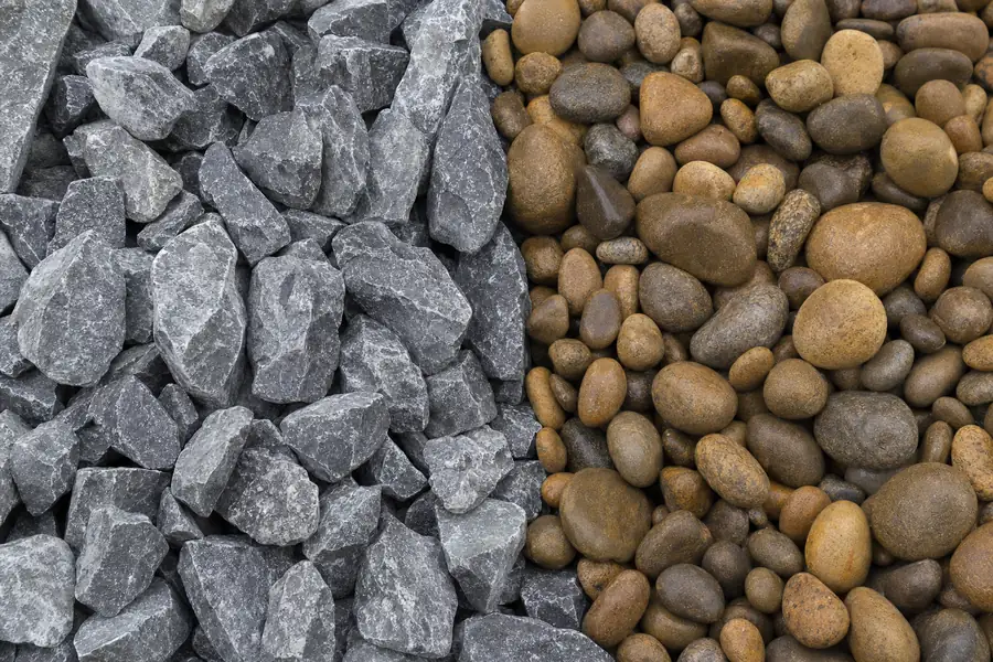 Macro photo pairing in one frame: the left half filled with sharp-edged gray crushed stone freshly fractured with visible dust on the facets, the right half with smooth water-worn river pebbles in warm browns and tans, a crisp dividing line down the middle, shallow depth of field, strong texture contrast that makes angular versus rounded instantly readable