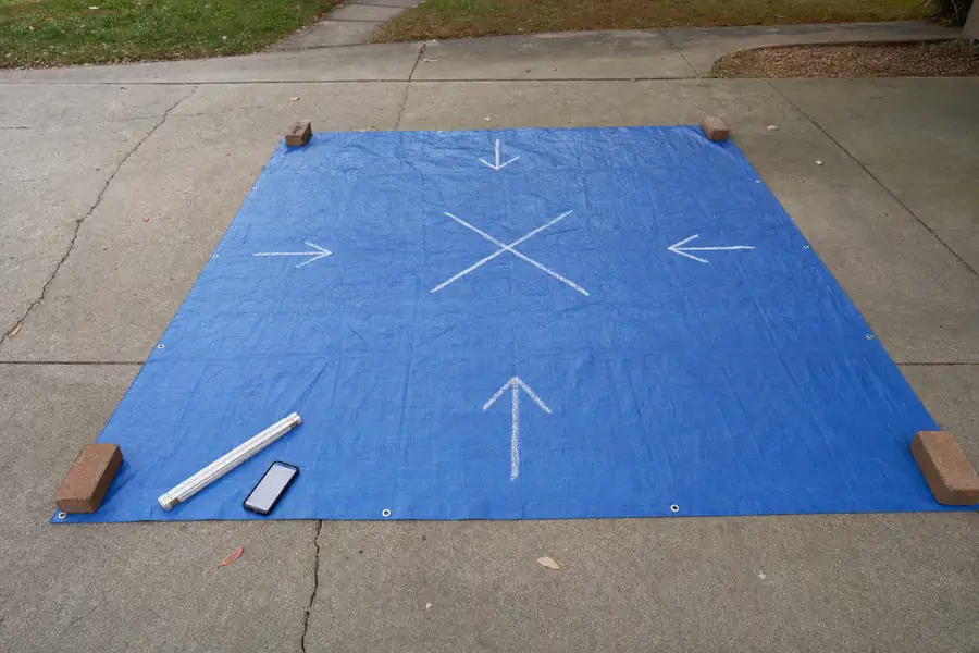 Detail photo of delivery-day preparation: a heavy blue tarp spread flat on a concrete driveway with bricks anchoring each corner, chalk arrows and a hand-drawn X marking the drop zone, a folding rule and phone with the order screen lying on the tarp edge, overcast even light, practical checklist-style clarity