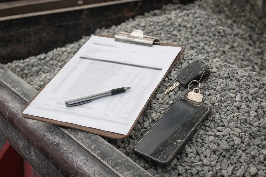 Detail photo of a printed supplier invoice on a clipboard resting on a small pile of number 57 stone: a pen, truck keys and a dusty phone beside it, line items softly out of focus, warm workshop light from the side, trust-and-transparency mood for a pricing article without any readable text
