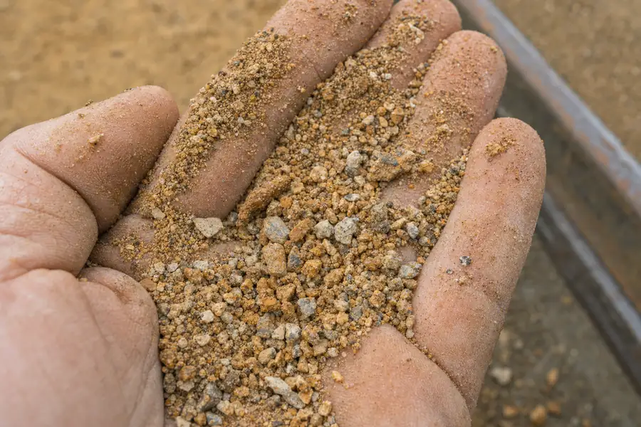 Extreme of decomposed granite texture in an open palm: coarse angular granite grains in gold, rust and gray with visible fines coating the fingers, a soft-focus DG path and steel edge in the background, shallow depth of field, tactile and material-honest