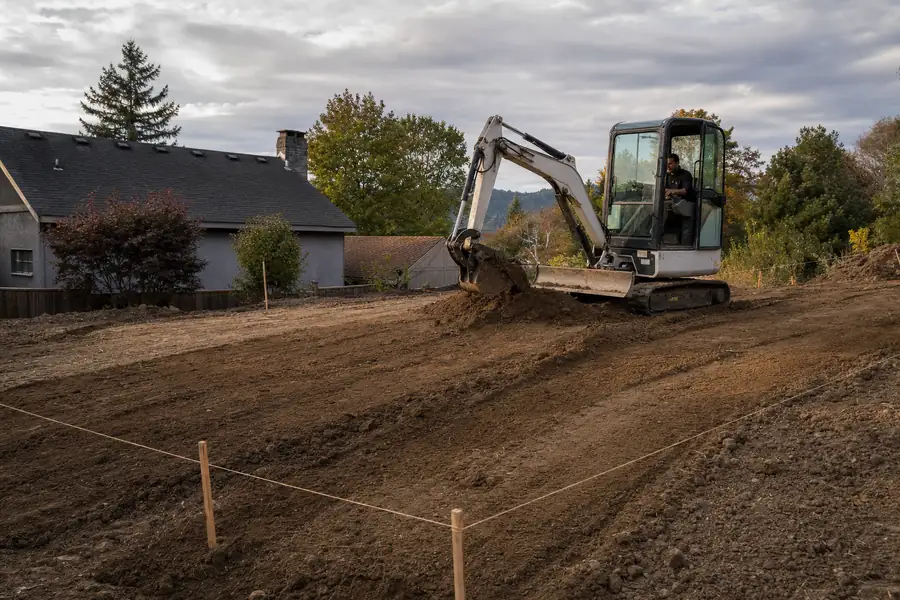 Photo of a compact excavator spreading and grading fill dirt across a sloped backyard being leveled: fresh cut lines in the soil, stakes with string marking final grade, operator focused, low evening sun raking across the graded surface to show contours, honest construction-progress feel