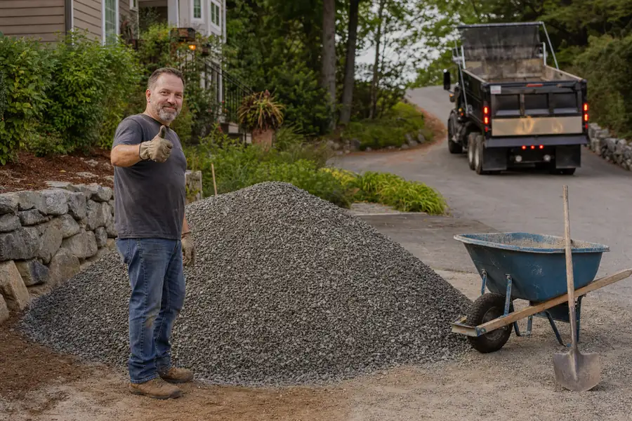 Warm photo of the moment after delivery: a homeowner in gloves giving a thumbs-up beside a fresh cone-shaped pile of 3/4-inch gravel while the empty dump truck pulls away down the street, wheelbarrow and shovel staged ready, late-afternoon golden light, satisfying job-started feeling with realistic residential detail