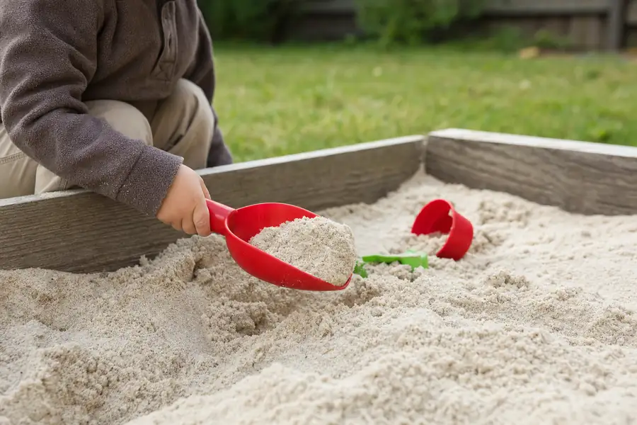 Photo of a backyard sandbox moment: a toddler's hands scooping clean bright play sand with a red plastic shovel, wooden sandbox frame and lawn softly blurred, gentle afternoon light, safety-and-quality warmth for the playground section