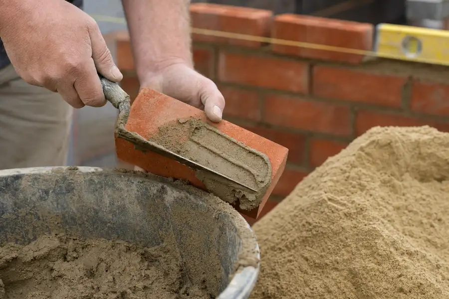 Photo of a mason's hands buttering a brick with mortar beside a mixing tub and a cone of fine mason sand, trowel mid-stroke, string line and level in the background, warm workshop light, the right-sand-for-the-job story told through craft detail